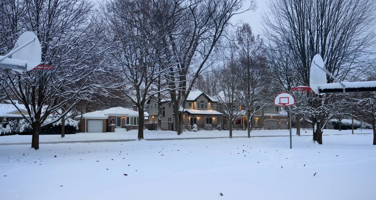 A snow-covered residential street at dusk with porch lights and windows lit in modest single-family homes