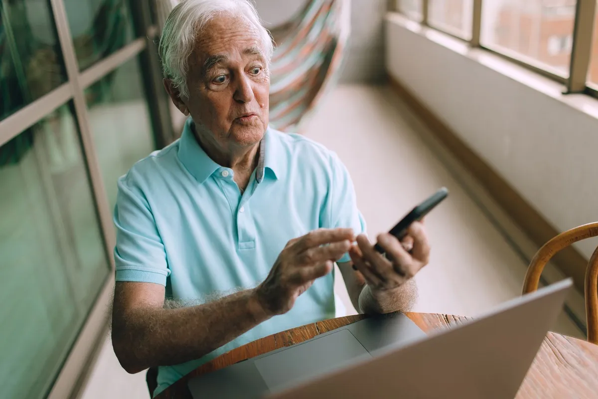 An older man in a light blue shirt sits at a table with a laptop open, holding his phone