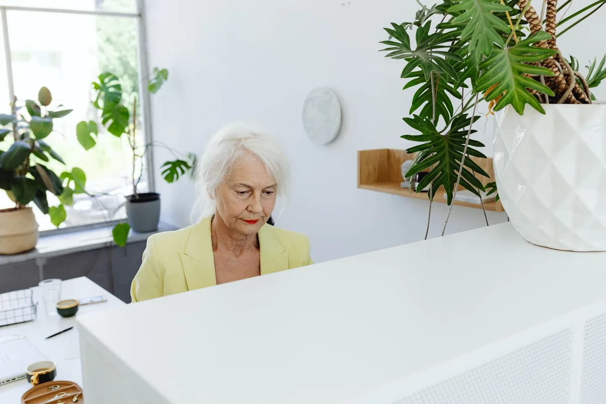 An older woman working at a desk in a bright, plant-filled office