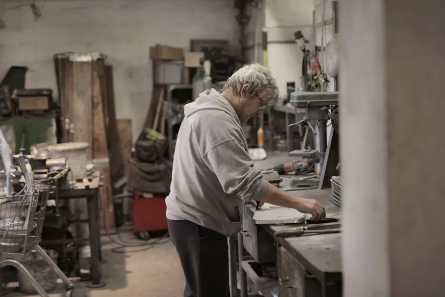An older worker at a workbench in a repair shop, focused on the piece in front of them