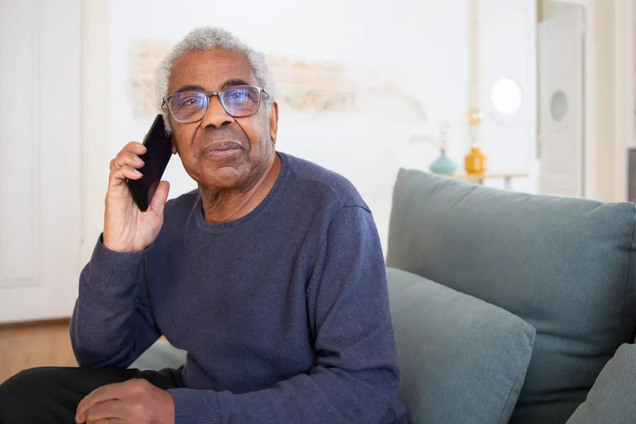 An older man with glasses sits on a sofa at home and listens on a cell phone