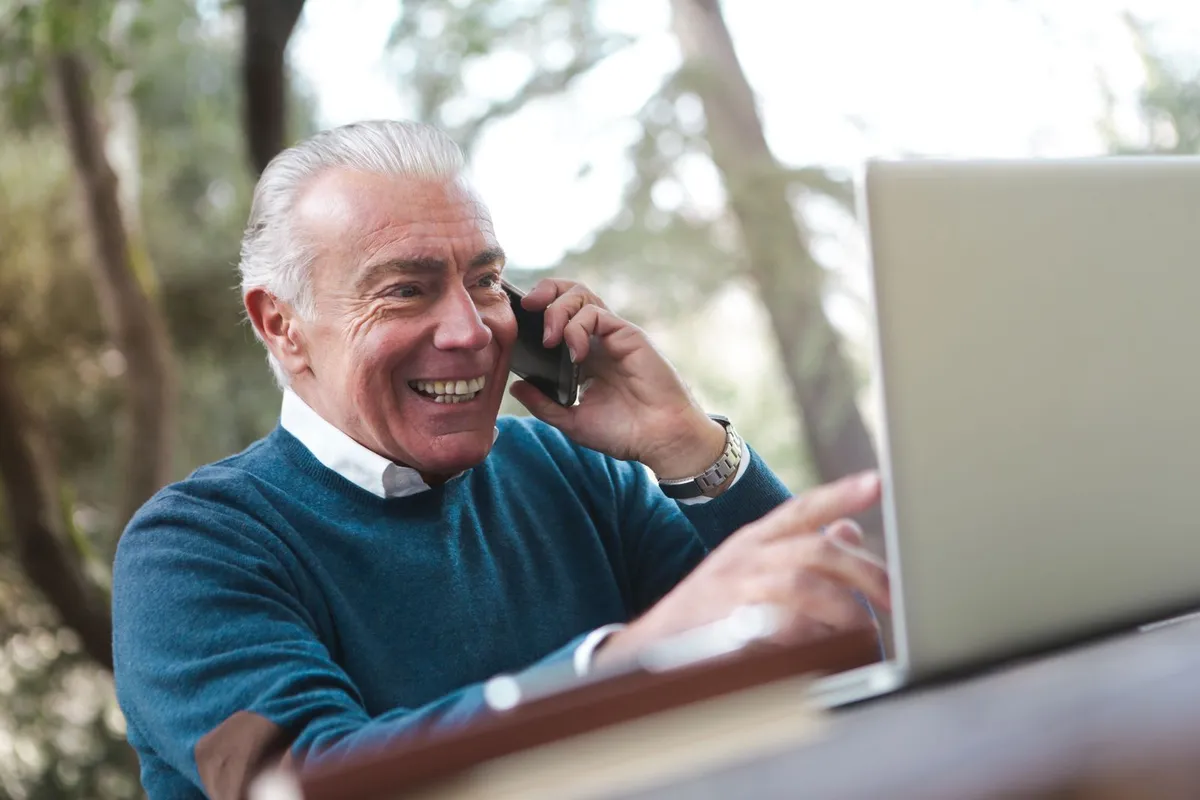 A smiling older man in a blue sweater talks on a cell phone while pointing at a laptop at an outdoor table