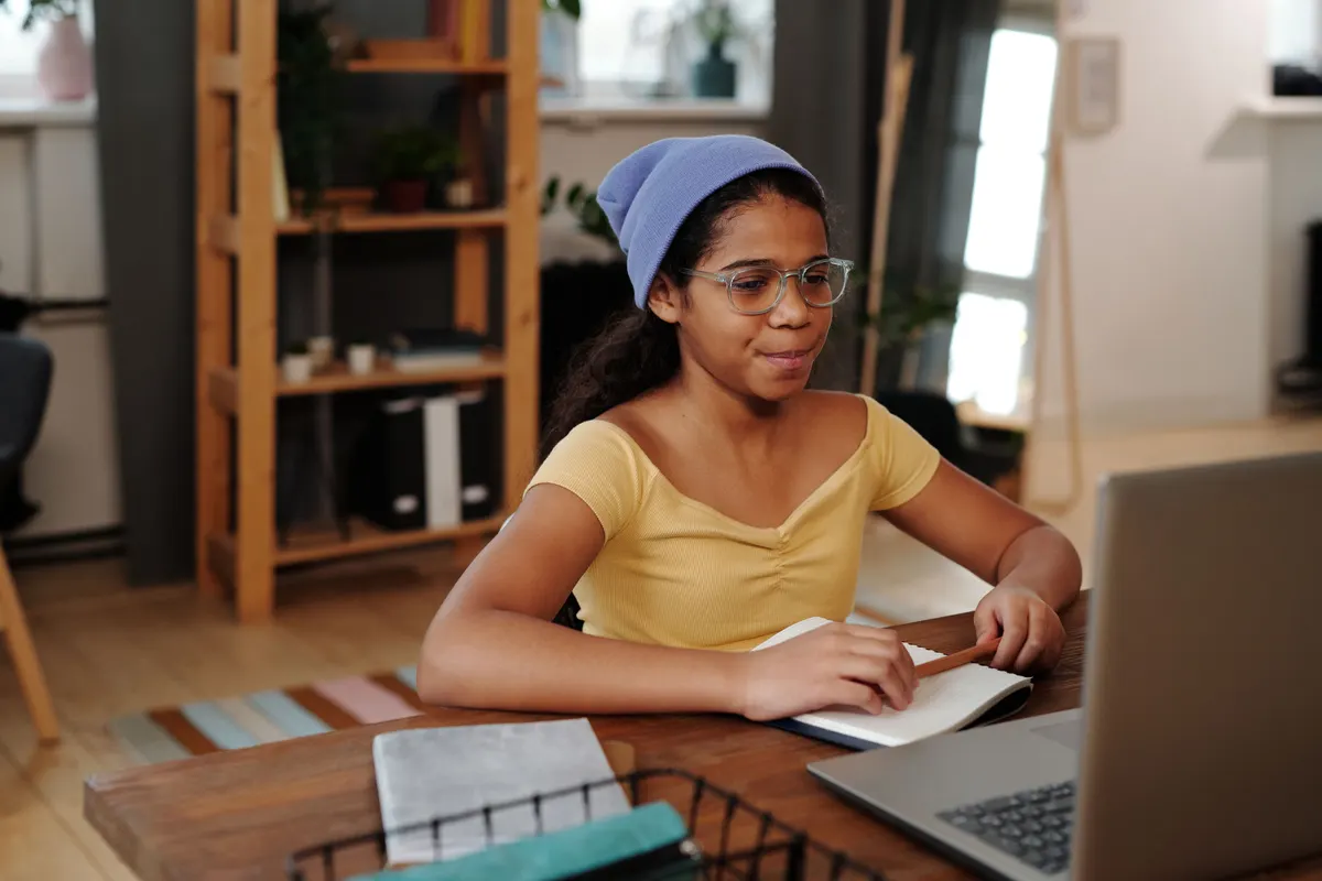 A girl in glasses and a blue knit hat sits at a wooden table writing in a notebook, with an open laptop beside her