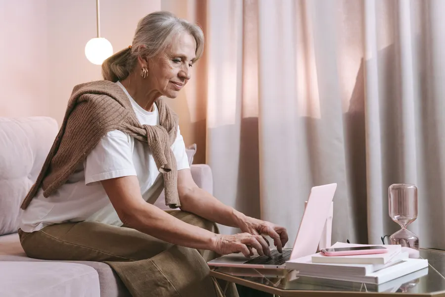 A gray-haired woman sits on a sofa and types on a laptop resting on a glass side table