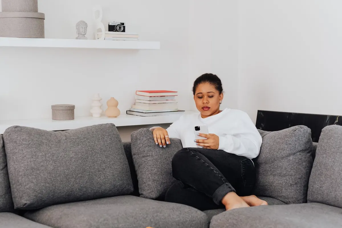 A woman in a white sweater sits on a gray couch at home, looking at her phone