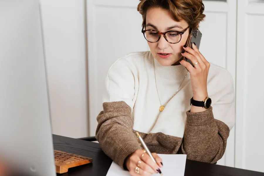 A woman at a desk takes notes on paper while talking on her phone