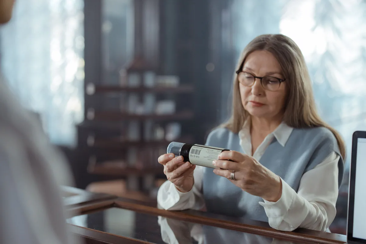 A senior woman with eyeglasses reads the label on a medicine bottle at a pharmacy counter