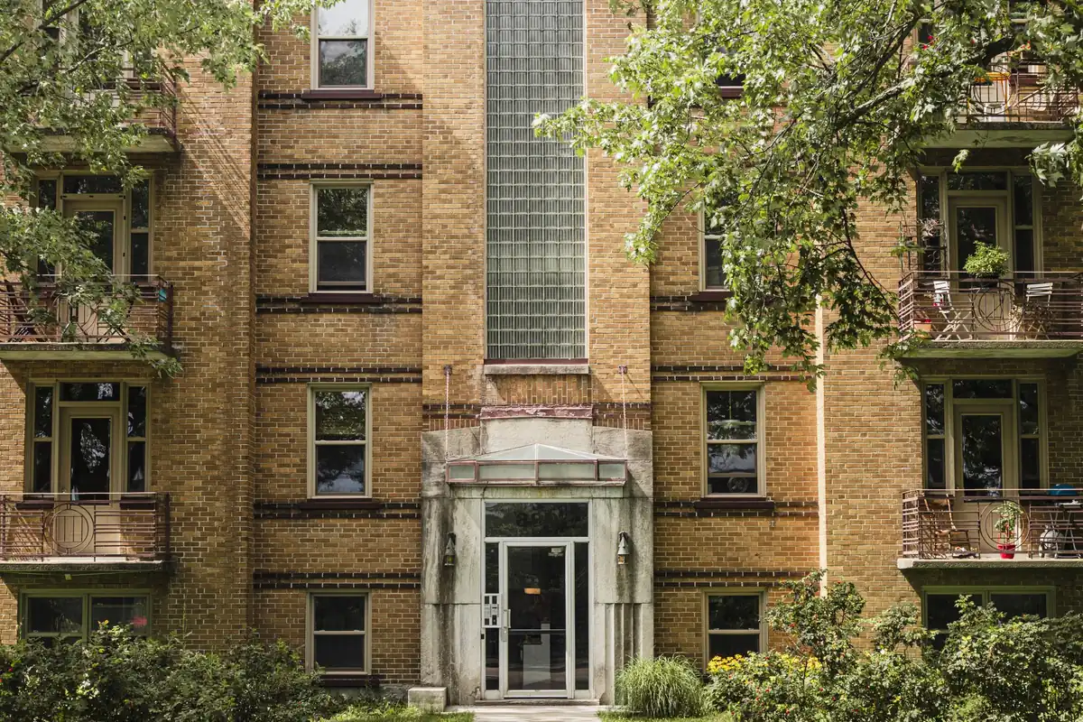 The front entrance of a classic brick apartment building framed by green trees on a city street