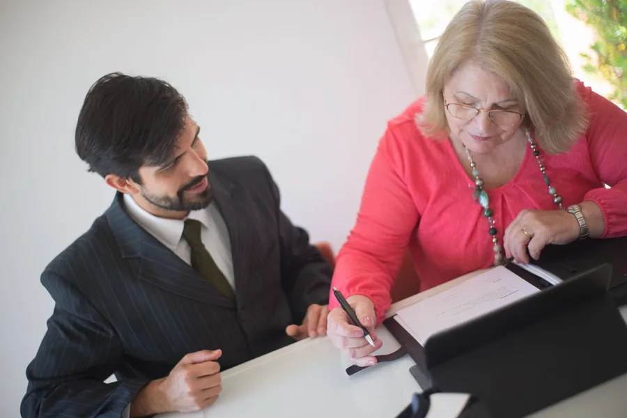 An older woman filling out an application form at a desk while a staff member helps her
