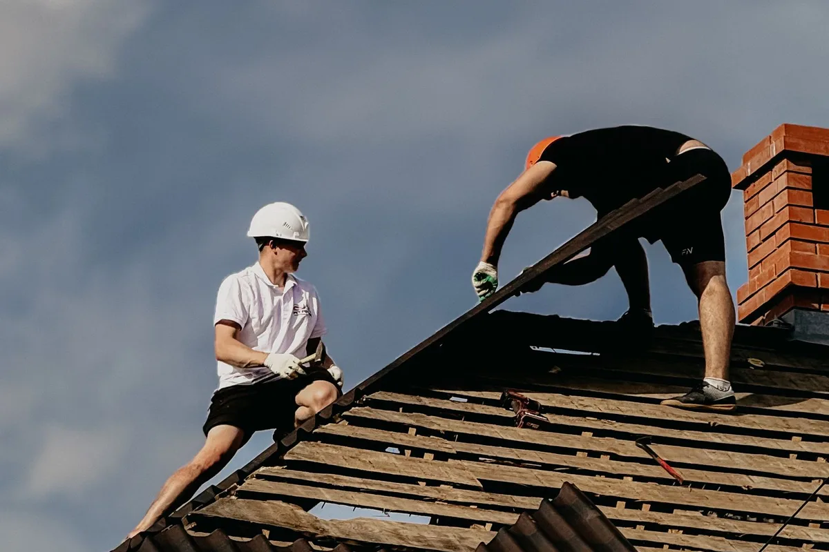 Two workers on a pitched roof lifting off old sheeting during a house repair
