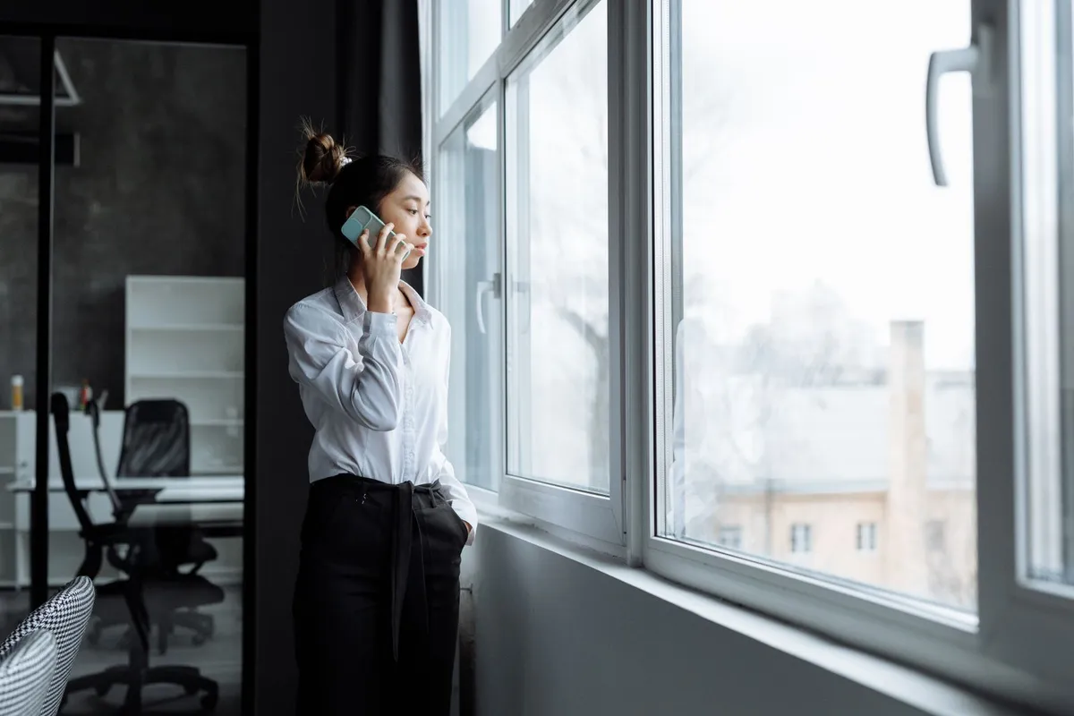 A woman stands by a large window talking on her phone