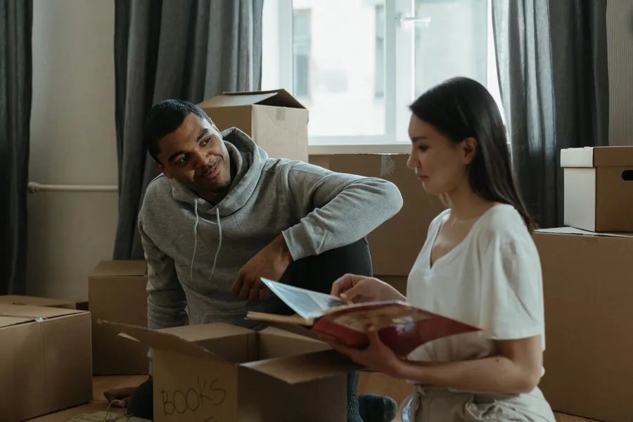 A couple sits among moving boxes in a bright room, one looking through a photo album