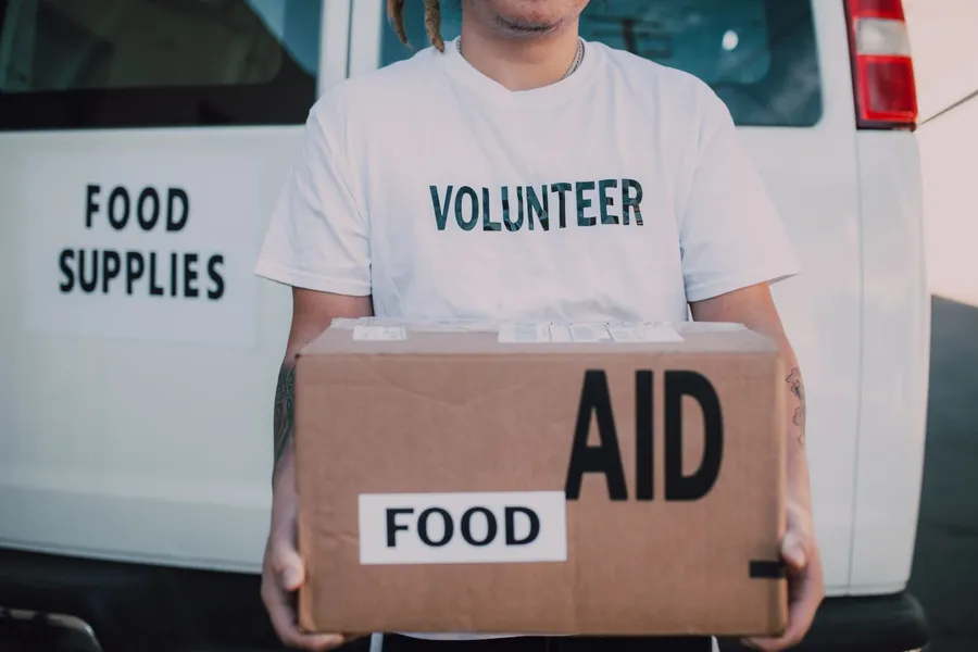 A volunteer holds a cardboard box of food aid in front of a white van