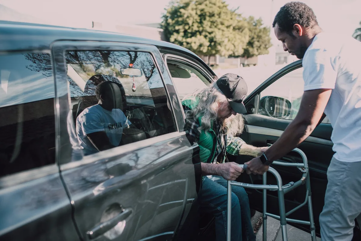 A younger man steadying an older man with a walker as he gets out of a car