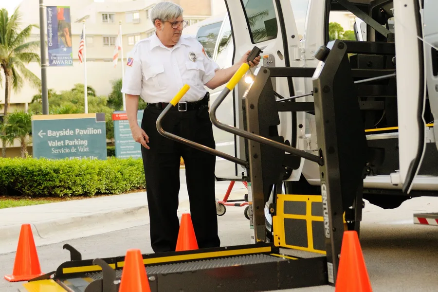 A uniformed transport aide lowering the wheelchair lift on a medical transport van outside a hospital entrance