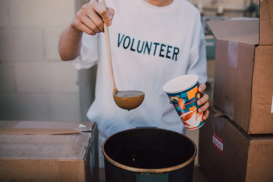 A volunteer in a "VOLUNTEER" shirt ladling hot soup into a cup beside stacked supply boxes