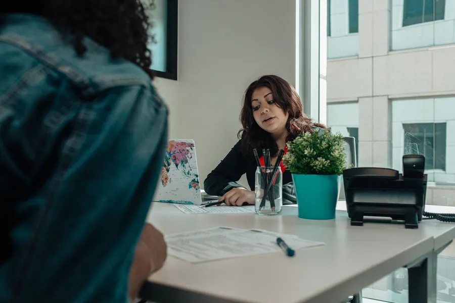 A person sitting across a desk from a staff member, with a printed application form and a pen on the desk between them