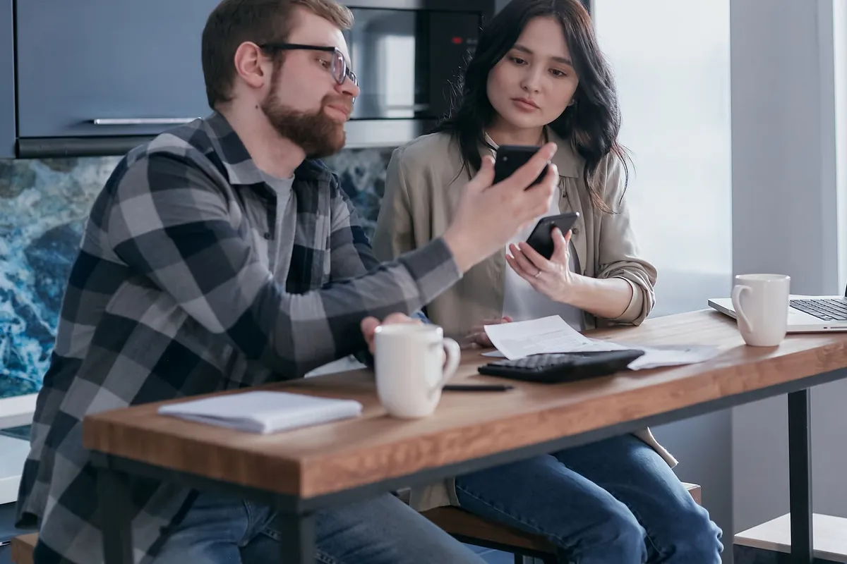 A couple sitting at a kitchen table, both looking at a phone, with a paper bill and a calculator in front of them