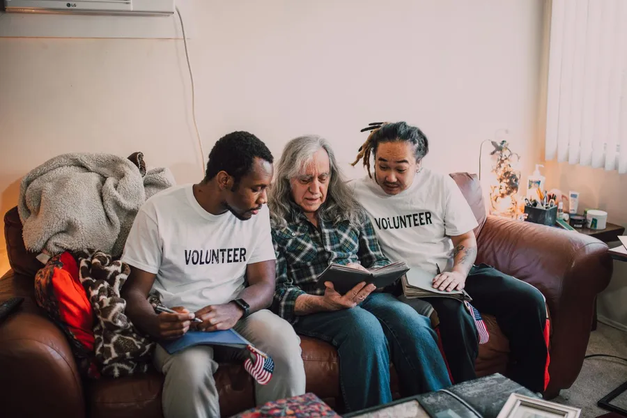 Two people in VOLUNTEER shirts sitting on a sofa in someone's living room, taking notes on a clipboard while the resident talks
