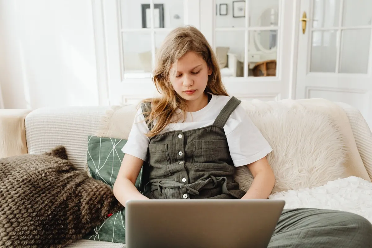 A teenager sits on a sofa at home with a laptop on her lap, looking down at the screen