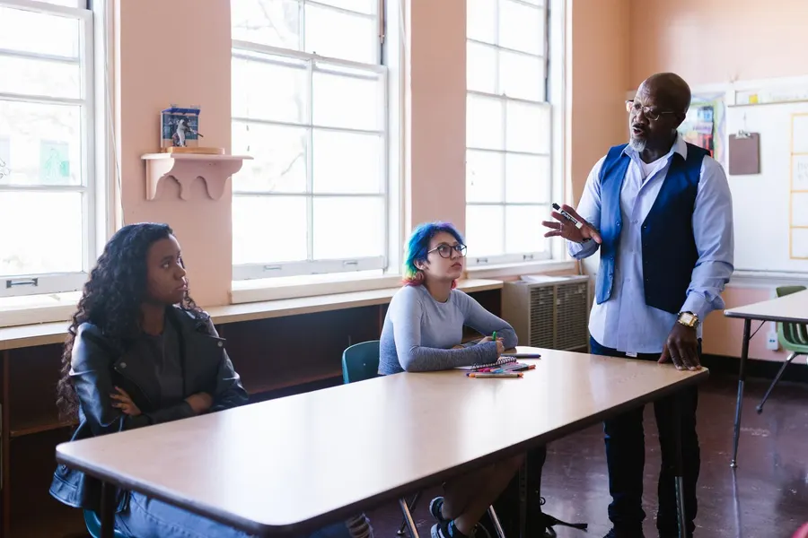 A school staff member standing at the end of a table talks with two high school students seated in a bright classroom
