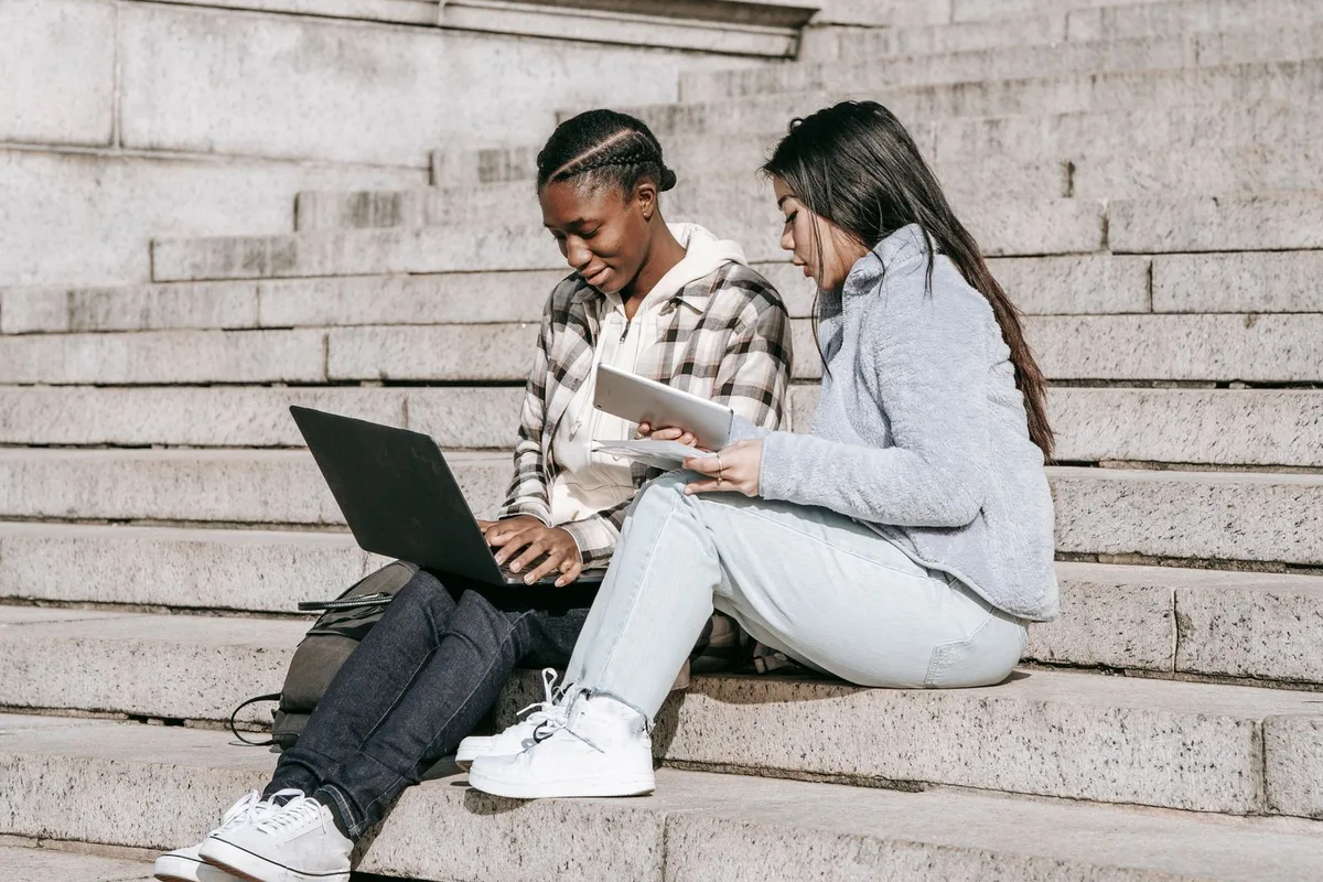 Two college students sitting on campus steps sharing a laptop and a tablet