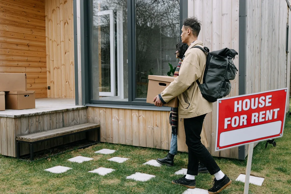 A young couple carrying moving boxes into a rental house with a "House for Rent" sign in the yard