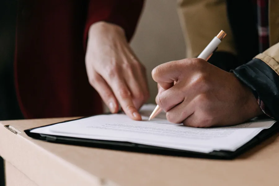 Two people signing a lease on a clipboard resting on a moving box inside an apartment