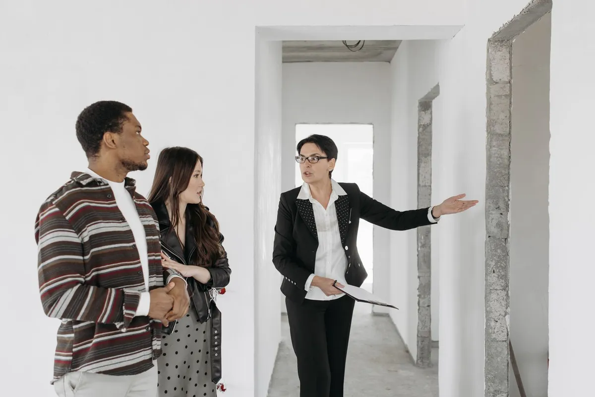 A man in a striped shirt and a woman with long dark hair listen as a woman with glasses and a clipboard points to a doorway in an empty, freshly painted apartment
