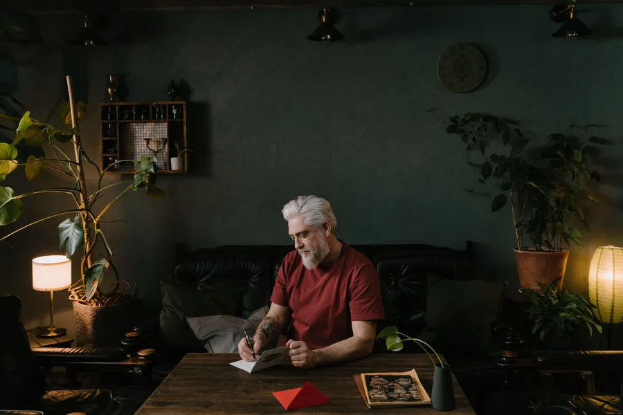 A man with white hair and a beard writes on a folded card at a wooden table in a dim, plant-filled room