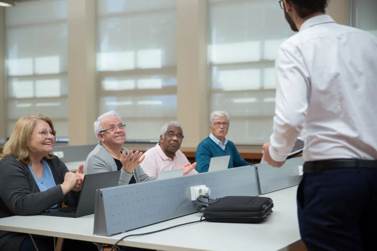 Four older adults sitting at a classroom table with laptops, smiling and talking with a teacher at the front of the room
