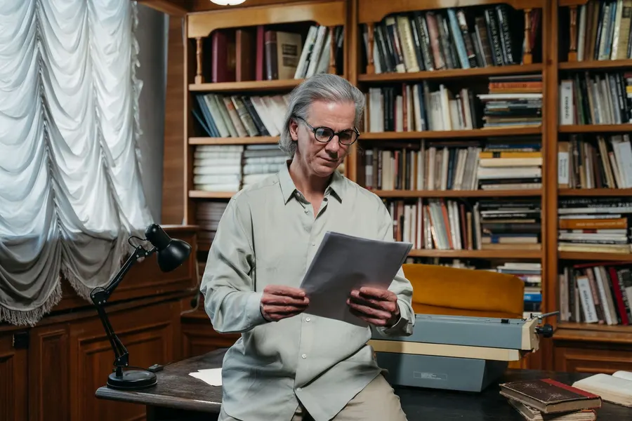 An older man with gray hair and round glasses reads a stack of printed pages while leaning on a desk in a room full of bookshelves