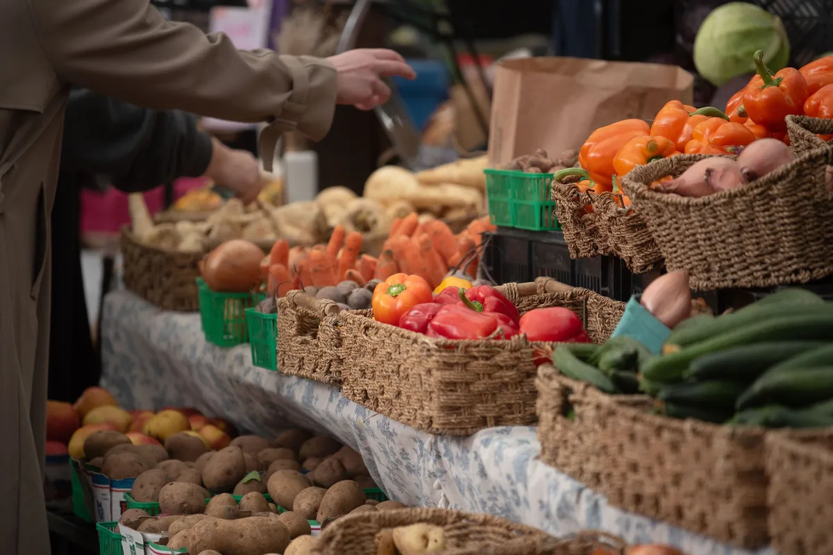 Baskets of peppers, cucumbers, potatoes, carrots and apples on a farmers market table, with a shopper reaching toward the produce