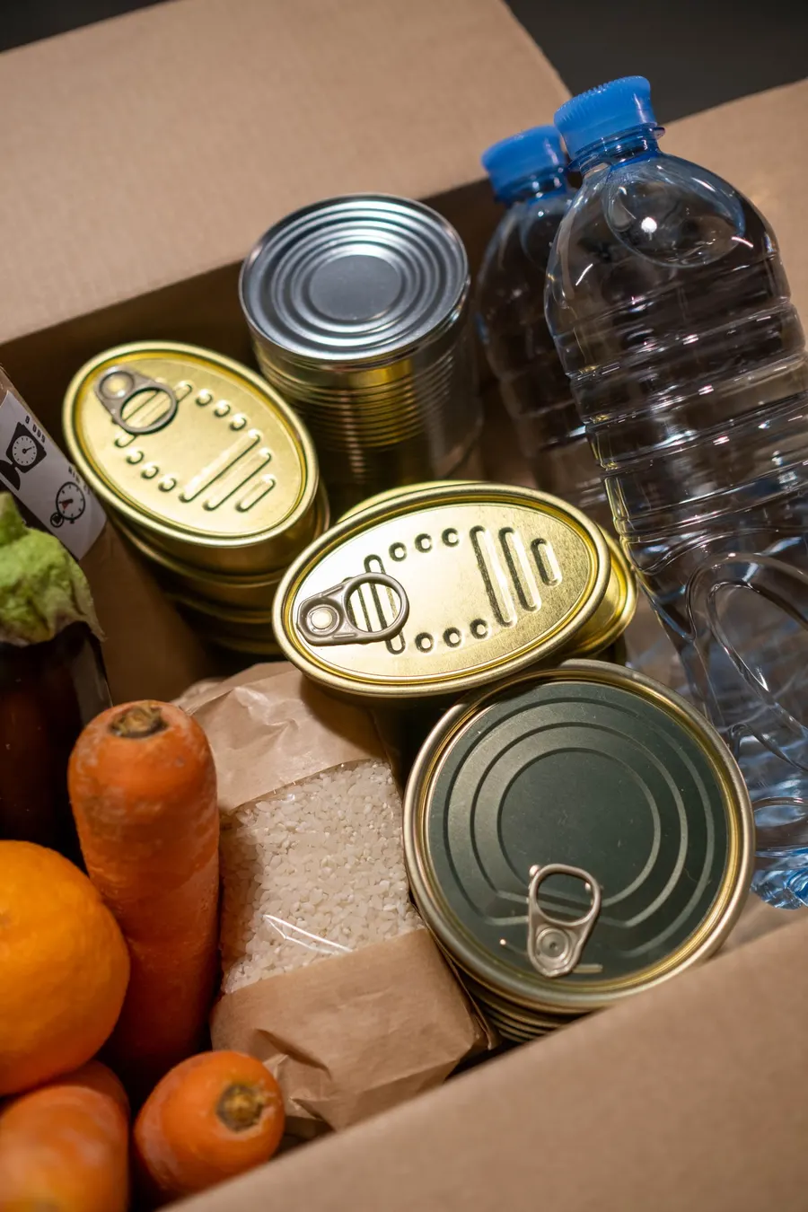 A cardboard food box packed with canned goods, a bag of rice, bottled water, carrots and oranges
