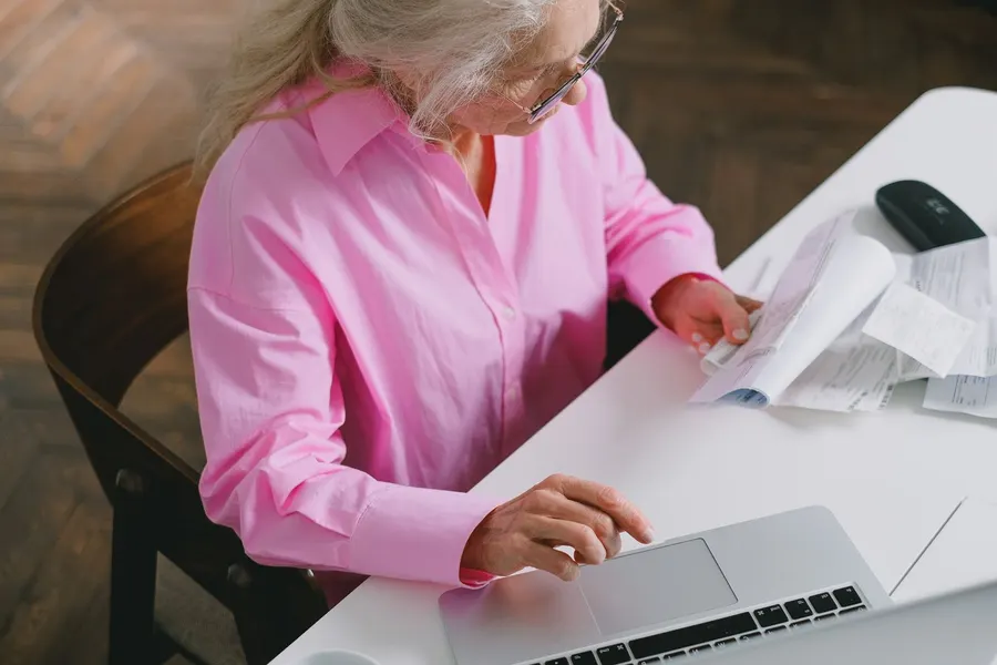 An older woman at a white desk sorting through statements and receipts next to an open laptop