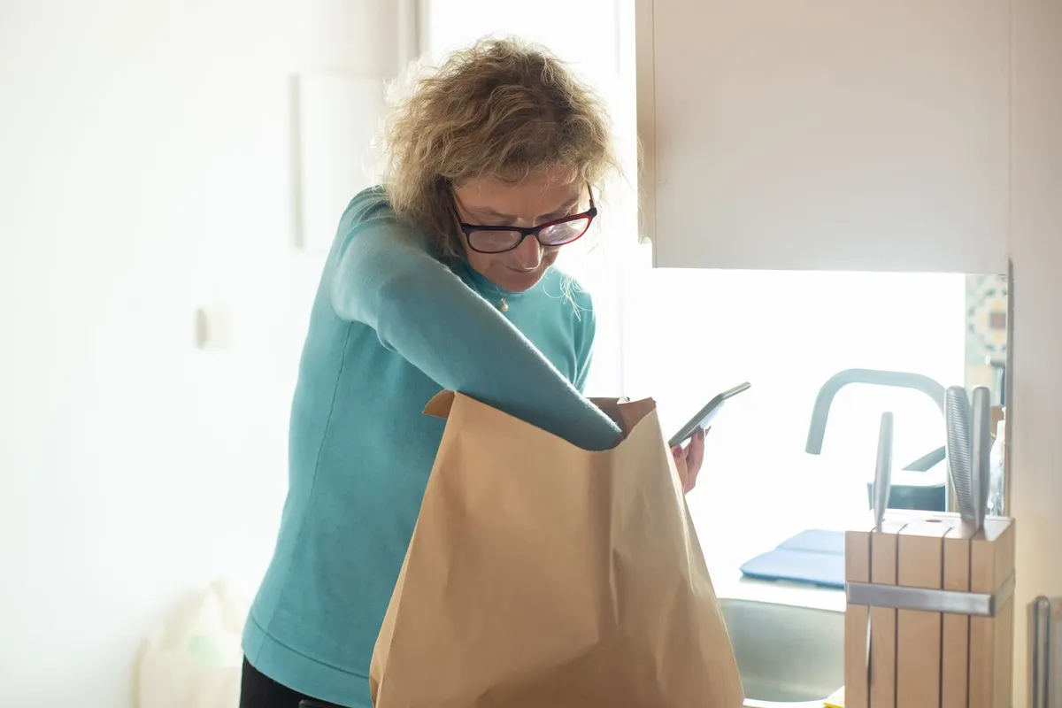 A woman in glasses and a teal sweater unpacking a paper grocery bag in a bright kitchen while checking her phone