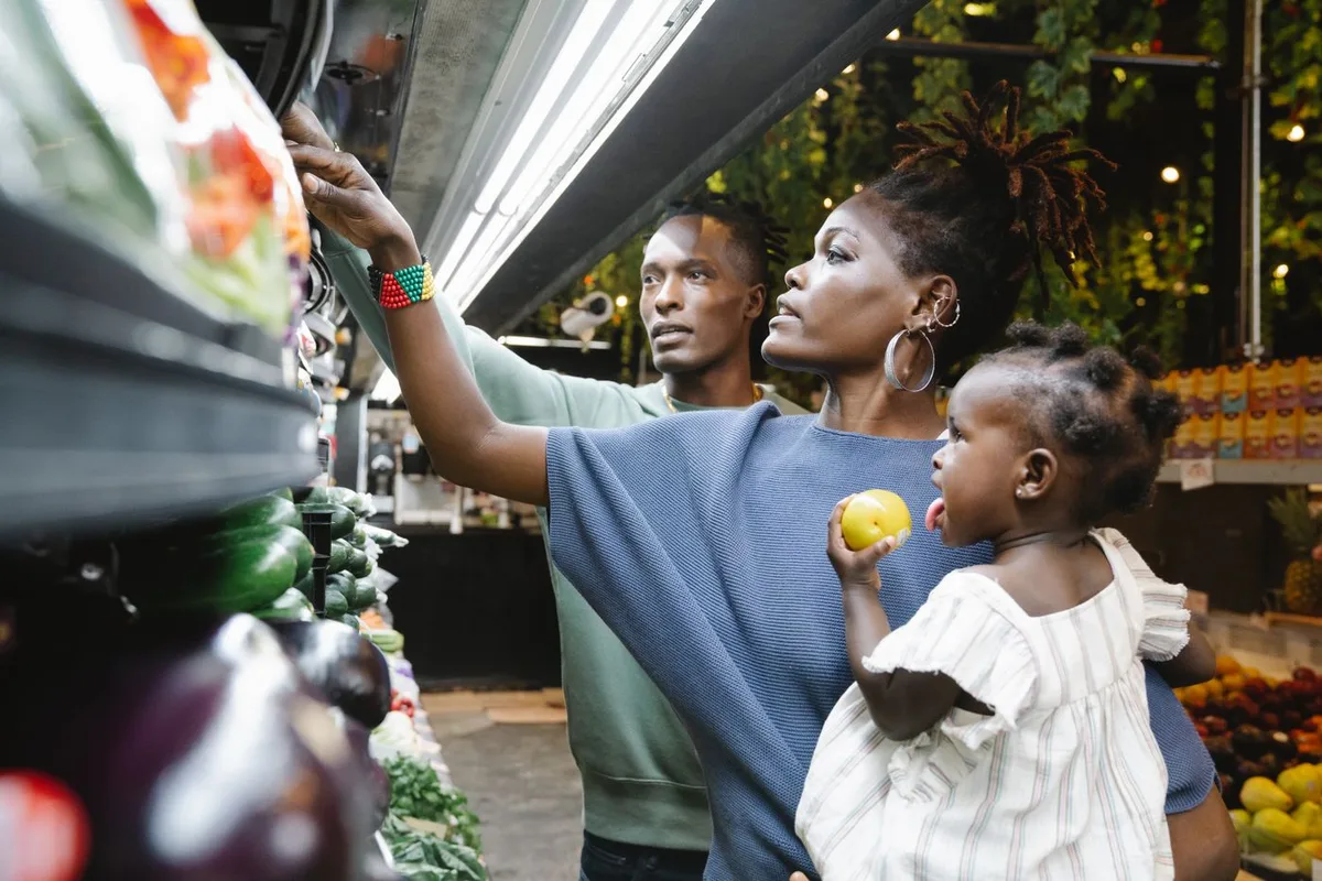 A family choosing fresh produce together at a grocery store, the parent reaching for a packaged salad while the child holds a lemon