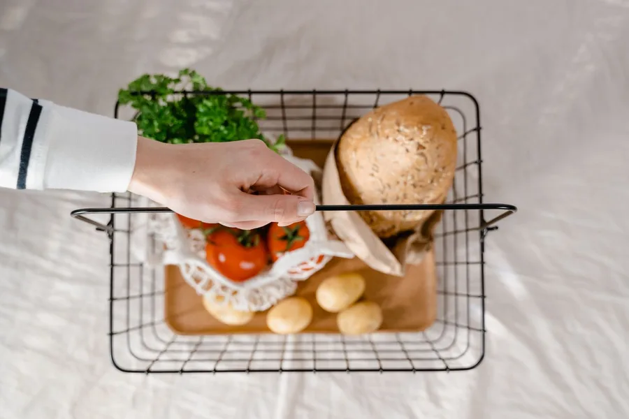 A hand carrying a wire shopping basket holding a loaf of bread, tomatoes, potatoes and fresh parsley