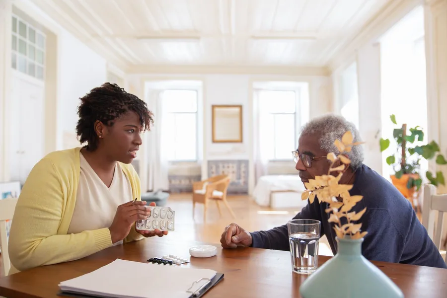 A home health worker and an older man at a dining table going over blister packs and a pill organizer, with a clipboard of paperwork on the table