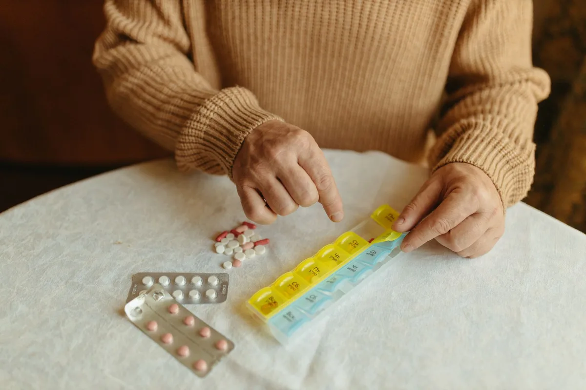 An older person's hands sorting tablets from blister packs into a weekly pill organizer on a kitchen table