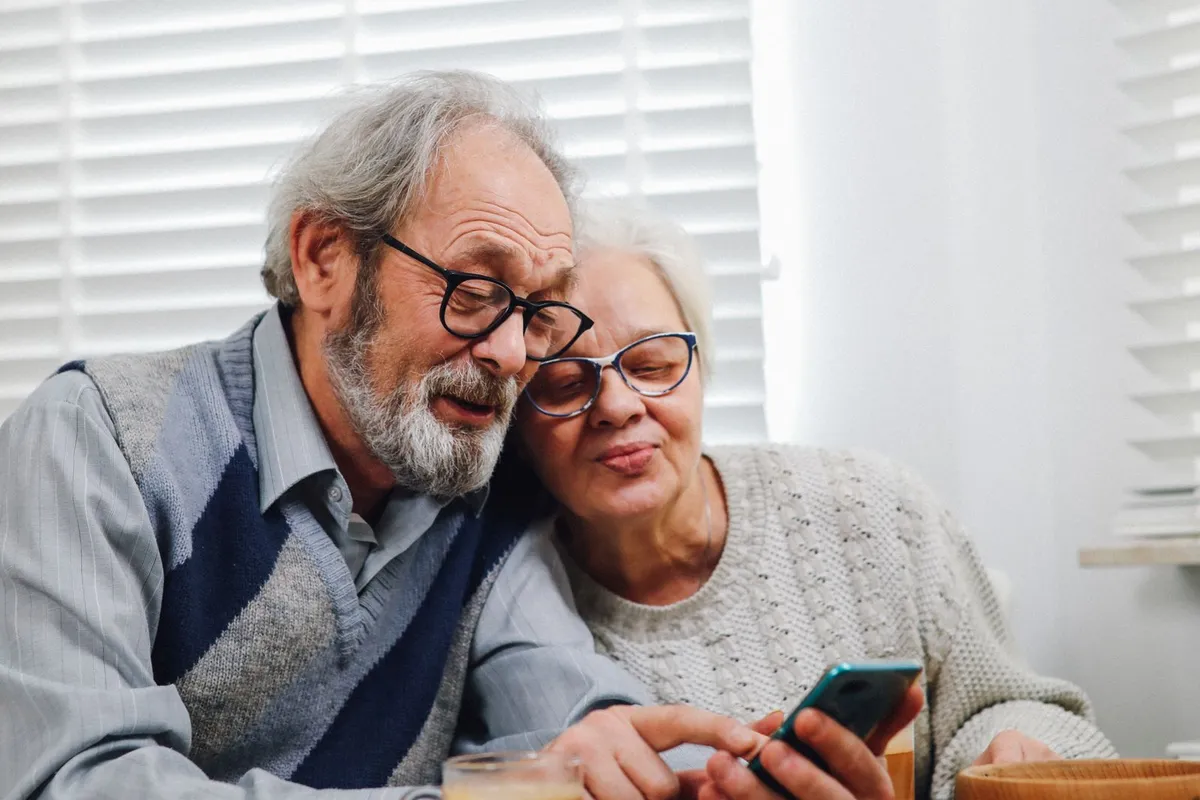 An older man with a gray beard and an older woman lean their heads together and smile while looking at a phone at a kitchen table