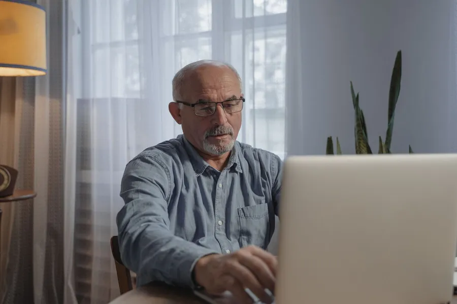 An older man in glasses and a denim shirt works on a laptop at a table by a bright window