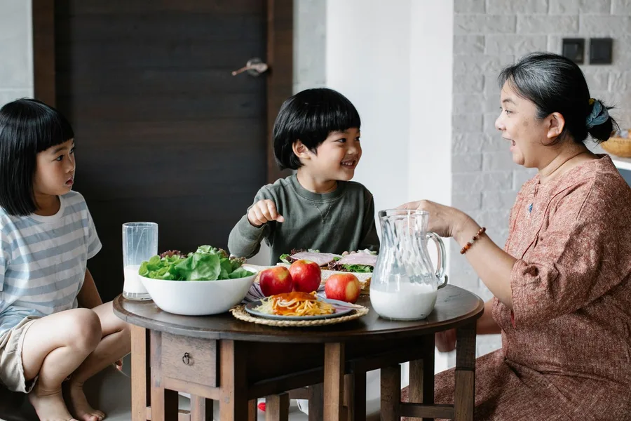 A grandmother pouring milk at the lunch table for two grandchildren, with salad and fruit on the table