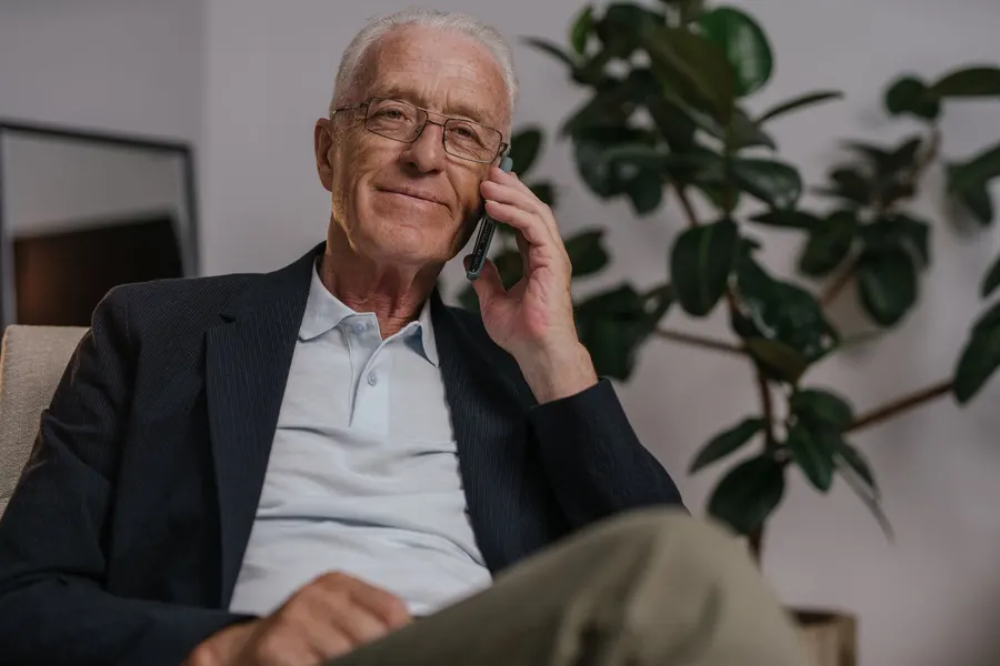An older man with white hair and glasses sits in an armchair at home, talking on a cell phone beside a large houseplant