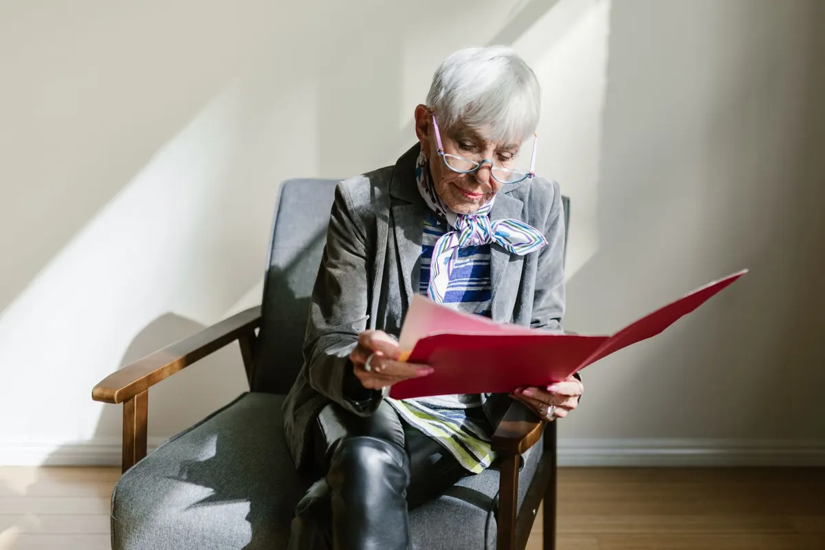 An older woman with short white hair and reading glasses sits in an armchair, reading papers in a red folder