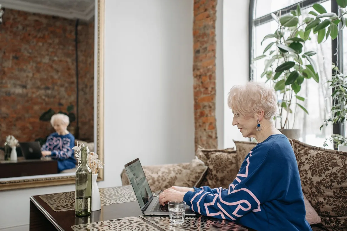 An older woman with short white hair sits at a table in a sunlit room, typing on a laptop