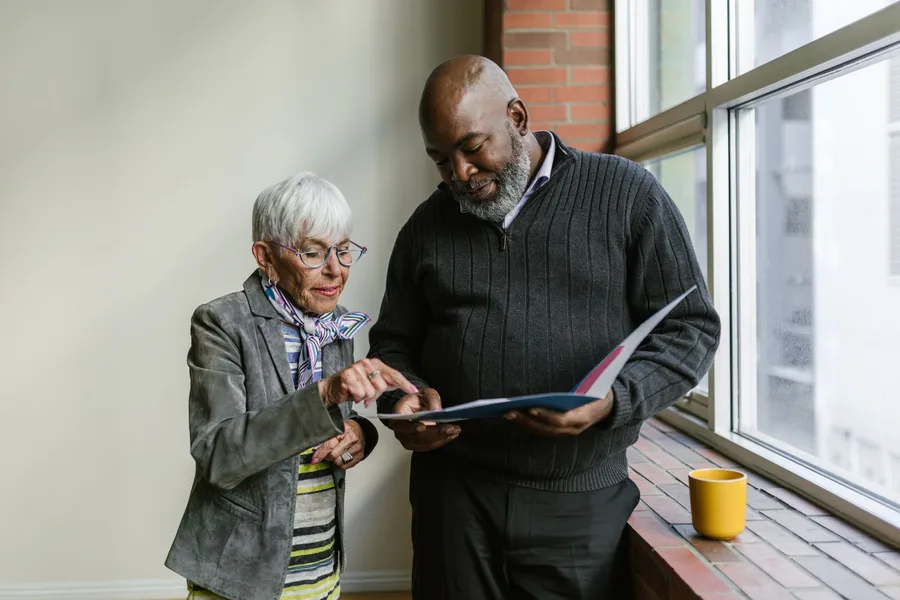 An older woman and a man stand by a window, going through the pages of an open folder together