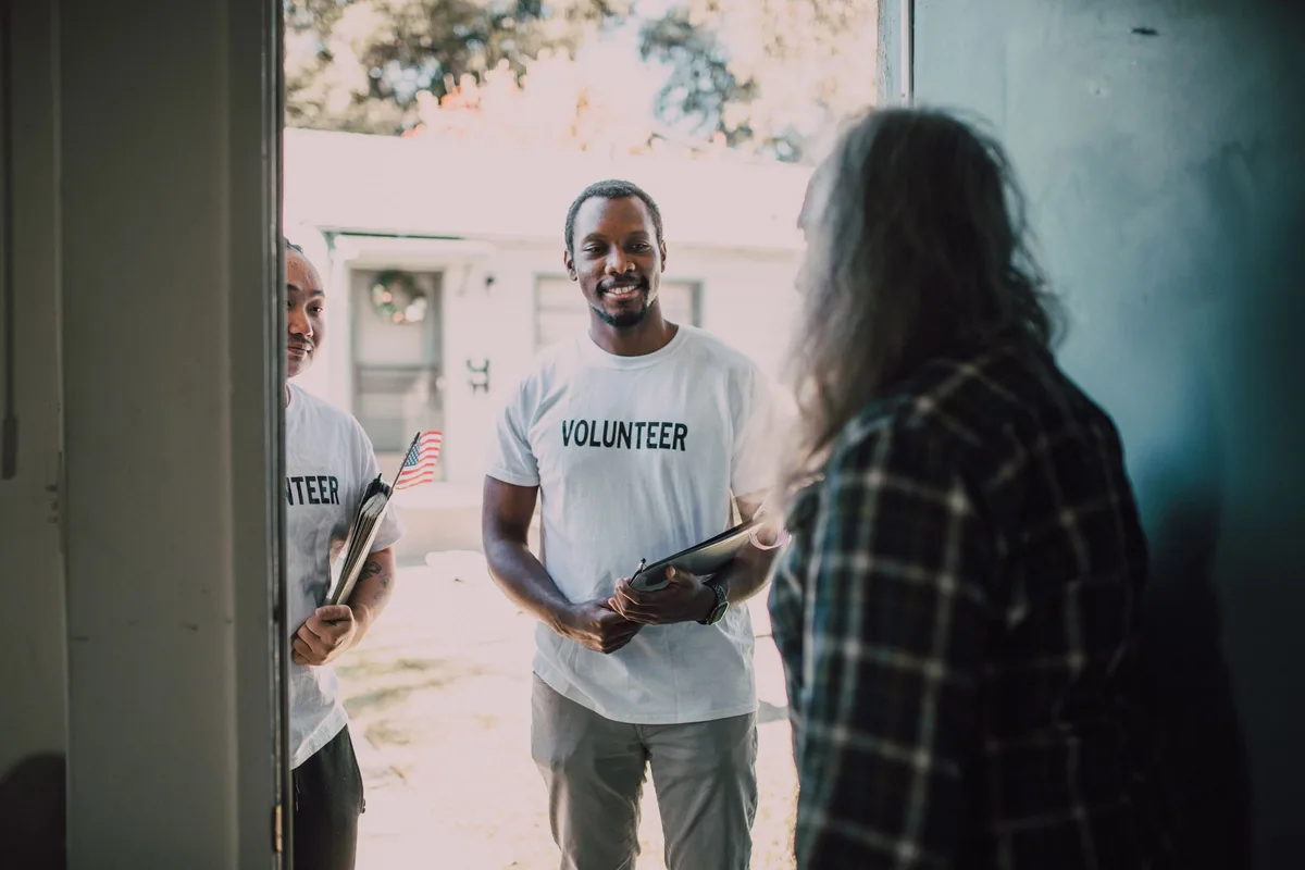 Two volunteers in "VOLUNTEER" shirts stand at a home's front door holding clipboards while the resident answers