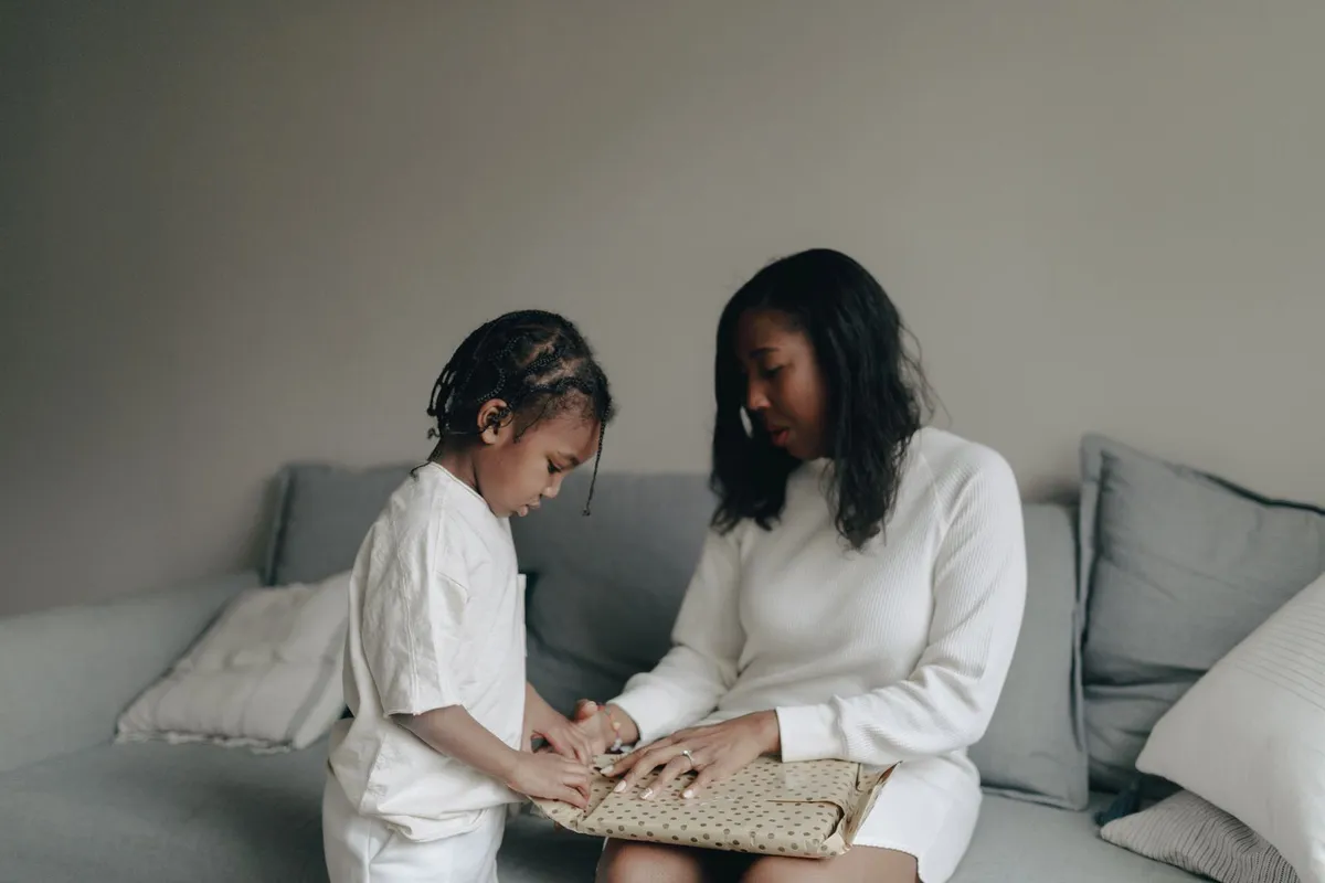 A mother sitting on a gray sofa helps her young son with a gift wrapped in polka-dot paper