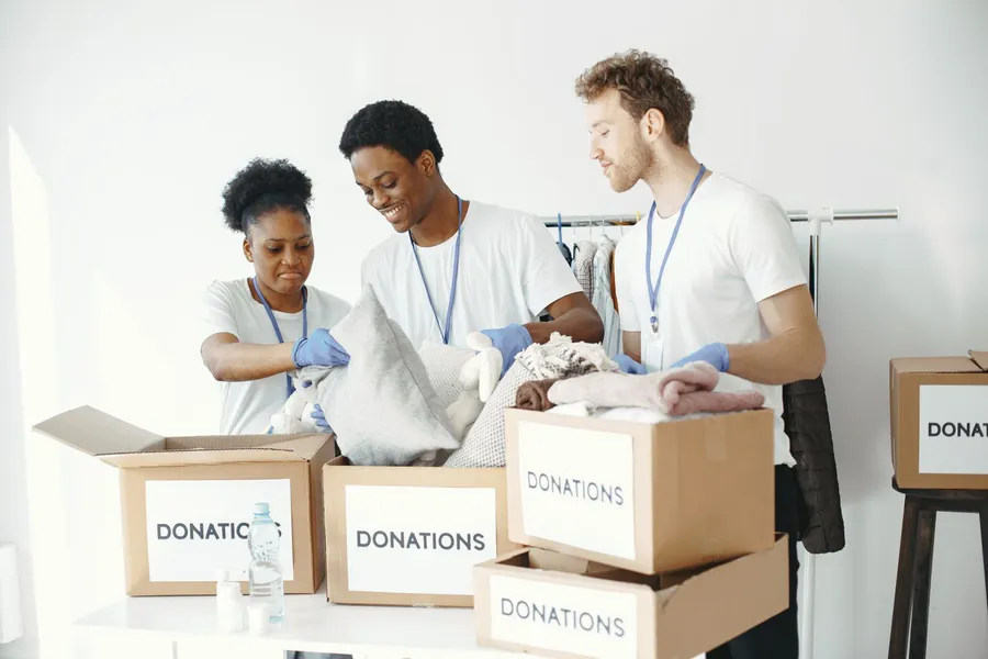 Three volunteers in matching white T-shirts and blue gloves sort donated items into cardboard boxes labeled donations